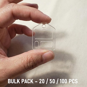 May include: A clear, rectangular plastic tag with a small hole at the top and a design of two connected rectangles. The text "BULK PACK - 20 / 50 / 100 PCS" is printed in white at the bottom of the image.