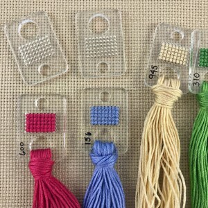 Pu&ograve; includere: Una collezione di schede colori per fili in plastica trasparente con piccoli campioni di colore cuciti e mazzi di filo attaccati in vari colori, tra cui rosa, blu, beige e verde. Ogni scheda ha un numero.