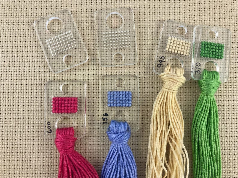 Pu&ograve; includere: Una collezione di schede colori per fili in plastica trasparente con piccoli campioni di colore cuciti e mazzi di filo attaccati in vari colori, tra cui rosa, blu, beige e verde. Ogni scheda ha un numero.