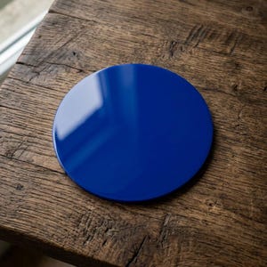 Peut inclure: Un dessous de verre rond et brillant, bleu cobalt, posé sur une surface en bois vieilli. Le dessous de verre reflète la lumière, soulignant sa finition lisse et polie. Un élément de décoration simple et élégant.