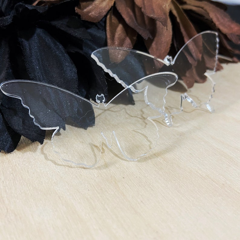May include: Two clear acrylic butterfly shapes on a light brown wooden surface. The butterflies are cut out and have a delicate, intricate design.