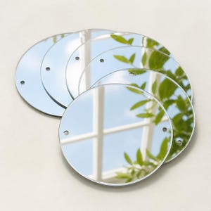 May include: A set of five circular, reflective mirror discs, each featuring two small, pre-drilled holes positioned on opposite sides. The smooth, silver-toned surfaces reflect a bright outdoor scene with a window frame and green foliage.