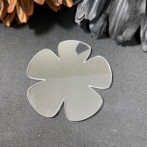 May include: A clear plastic flower-shaped cutout with five petals. The cutout is on a dark gray fabric background.