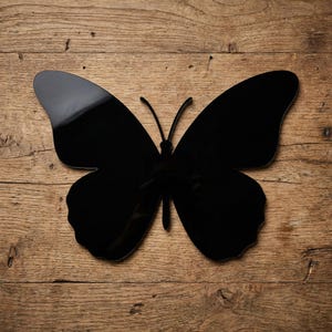 May include: A large, glossy black butterfly silhouette. The butterfly is centered on a wooden surface with visible grain. The wings are fully extended, and the antennae are thin and curved. The overall design is simple and elegant.