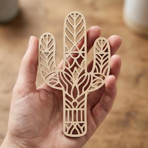 Könnte beinhalten: Eine lasergeschnittene Holzdekoration in Form eines Kaktus mit komplizierten geometrischen Mustern. Der hellbraune Kaktus wird in einer Hand gehalten, was das detaillierte Design hervorhebt. Der Kaktus hat eine dreidimensionale Form mit ausgeschnittenen Abschnitten.
