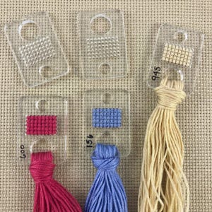 Pu&ograve; includere: Guide di cucitura in plastica trasparente con vari campioni di filo colorato. Le guide presentano piccoli motivi cuciti e sono etichettate con numeri. I fili sono rosa, blu e beige. Lo sfondo &egrave; un tessuto beige.