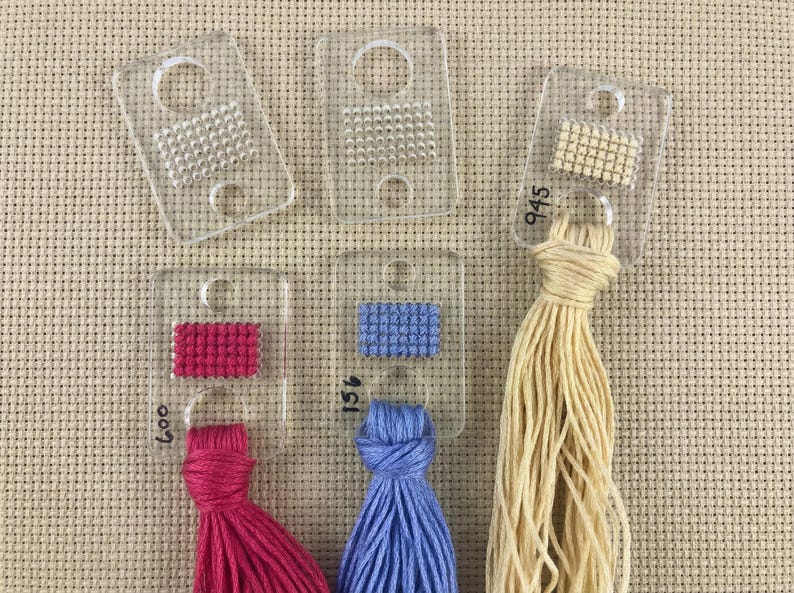 Pu&ograve; includere: Guide di cucitura in plastica trasparente con vari campioni di filo colorato. Le guide presentano piccoli motivi cuciti e sono etichettate con numeri. I fili sono rosa, blu e beige. Lo sfondo &egrave; un tessuto beige.