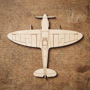 May include: A detailed wooden model of a Spitfire aircraft, seen from above. The light brown plane features etched lines defining the wings, fuselage, and propeller. It rests on a wooden surface, showcasing the aircraft's design.