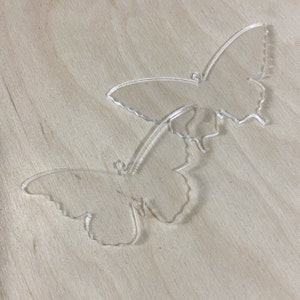 May include: Two clear acrylic butterfly shapes on a light brown wooden surface.