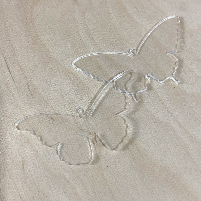 May include: Two clear acrylic butterfly shapes on a light brown wooden surface.