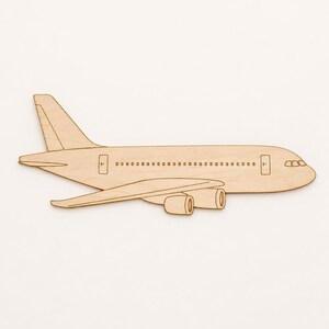 May include: A flat, light-colored wooden cutout shaped like a commercial passenger airplane. The design features a side profile of the aircraft, including the fuselage, wings, tail fin, and two jet engines mounted under the wings, with etched details.