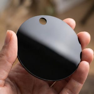 May include: A round, black, glossy object with a small hole at the top. The object is held in a person's hand. The surface reflects light, creating highlights. The item appears to be a decorative element or a component of a larger product.