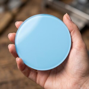 May include: A round, light blue glass coaster is held in a hand. The coaster has a smooth, reflective surface and a solid colour. The background is a blurred wooden surface.
