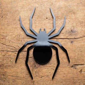 May include: A black, stylized spider decoration with eight legs and a rounded body. The spider is made of a glossy material and is set against a textured, wooden background. This decorative piece is suitable for Halloween.