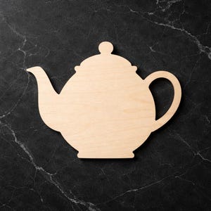 May include: A light-coloured wooden teapot cutout with a handle, spout, and lid. The teapot is set against a dark, marbled background. The cutout is likely for crafting or decorative purposes.