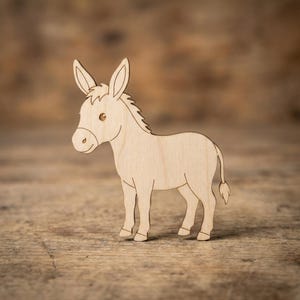 May include: A wooden donkey figurine, standing on a wooden surface. The donkey is light brown with a simple, cartoon-like design. It has large ears, a small tail, and is facing left. The background is a blurred, warm-toned wooden surface.