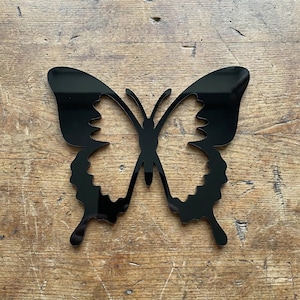 May include: A black butterfly-shaped decorative object. The butterfly has a symmetrical design with detailed wing edges. The object is made of a shiny, reflective material and is set against a wooden background.
