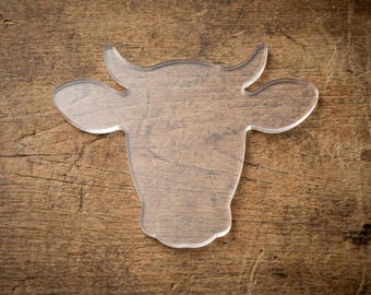 May include: A clear acrylic cow head shape, featuring horns and ears, is displayed on a textured wooden surface. The cow head is a transparent, decorative item, likely a coaster or a similar craft supply.