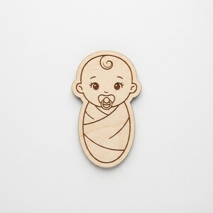 Puede incluir: Un recorte de madera con forma de bebé con detalles grabados. El bebé tiene un chupete, un cuerpo envuelto y un rizo de pelo rizado. El recorte es de color marrón claro y tiene un acabado liso. El bebé mide unos 10 cm de alto.