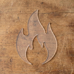 May include: A transparent, flame-shaped acrylic template, positioned on a wooden surface. The template's edges are clearly defined, forming a stylized fire design. The wood grain provides a warm, natural backdrop.