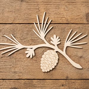 May include: A light-colored, flat wooden cutout shaped like a pine branch with needles and a single pinecone. The design features a central stem with three clusters of needle-like projections and two small, rounded leaf-like shapes near the pinecone.