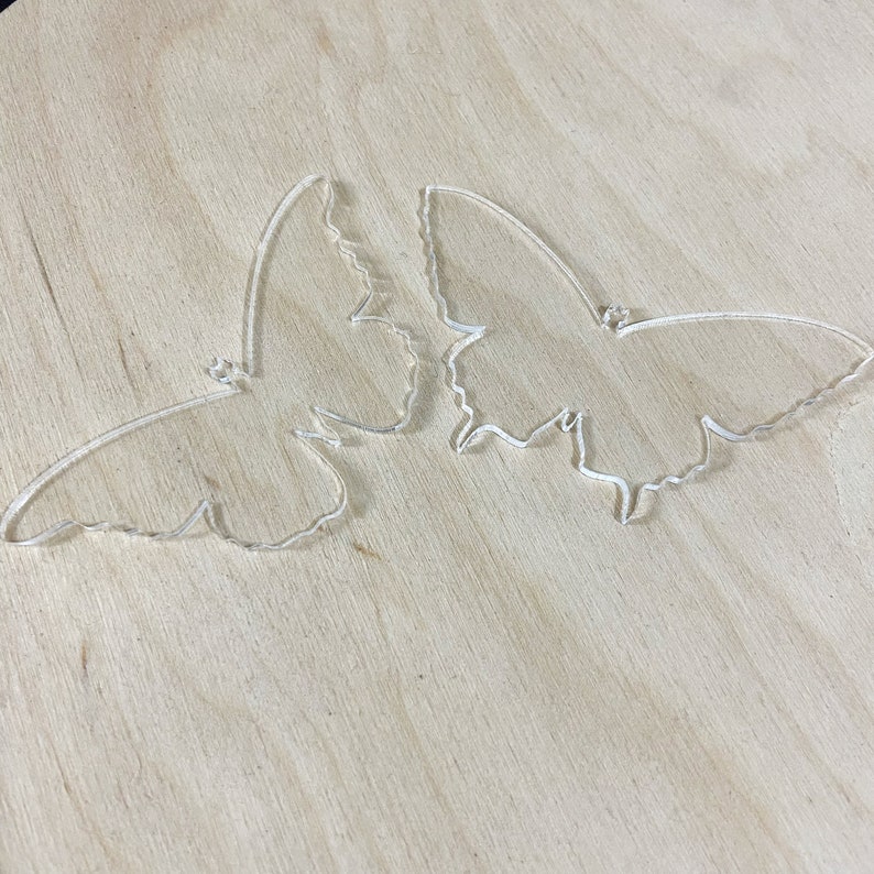 May include: Two clear acrylic butterfly shapes on a light brown wooden surface.