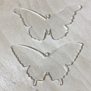 May include: Two clear acrylic butterfly shapes, one slightly larger than the other, on a light wood background.