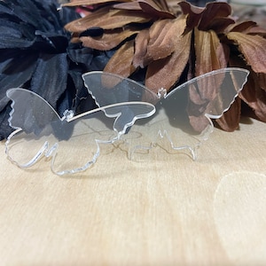 May include: Two clear acrylic butterfly shapes, one slightly larger than the other, on a light brown wooden surface.