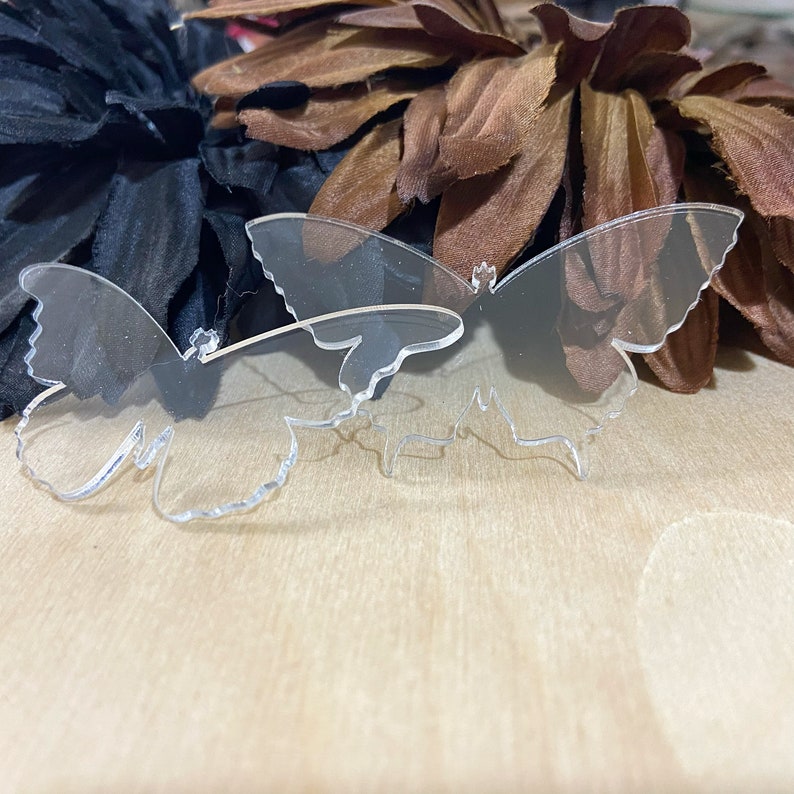 May include: Two clear acrylic butterfly shapes, one slightly larger than the other, on a light brown wooden surface.