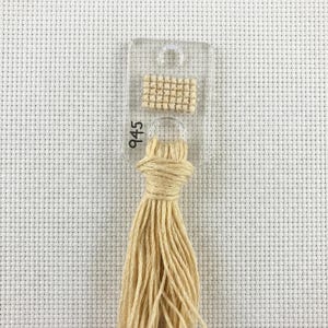 Pu&ograve; includere: Un campione di filo per punto croce di colore beige chiaro, contrassegnato con il numero 945. Il filo &egrave; attaccato a una scheda di plastica trasparente con un piccolo quadrato cucito per riferimento cromatico. Lo sfondo &egrave; un tessuto bianco a punto croce.