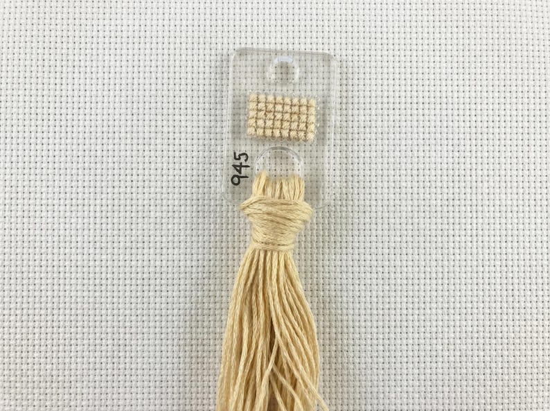 Pu&ograve; includere: Un campione di filo per punto croce di colore beige chiaro, contrassegnato con il numero 945. Il filo &egrave; attaccato a una scheda di plastica trasparente con un piccolo quadrato cucito per riferimento cromatico. Lo sfondo &egrave; un tessuto bianco a punto croce.