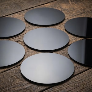 May include: A collection of seven black, round coasters with a glossy finish. The coasters are arranged on a rustic, wooden surface, showcasing their reflective quality and minimalist design. The coasters are a dark gray color.
