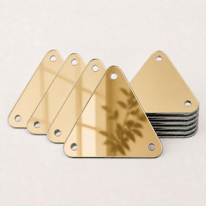 Op de afbeelding: Een stapel goudkleurige driehoekige acryl spiegelstukken. Elk stuk is voorzien van drie kleine ronde gaatjes, één in elke hoek, en heeft een sterk reflecterend, glad oppervlak dat de reflectie van een plant en licht toont.