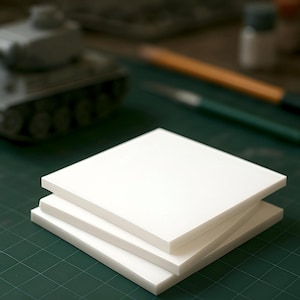 May include: Three white square pieces of foam board stacked on top of each other on a green cutting mat.