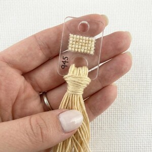 Pu&ograve; includere: Una scheda colori per fili in plastica trasparente con un piccolo campione di tessuto a punto croce beige e il numero 945 stampato sopra. Un fascio di filo da ricamo beige &egrave; attaccato alla scheda.