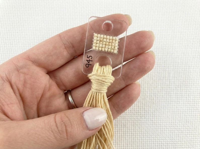 Pu&ograve; includere: Una scheda colori per fili in plastica trasparente con un piccolo campione di tessuto a punto croce beige e il numero 945 stampato sopra. Un fascio di filo da ricamo beige &egrave; attaccato alla scheda.