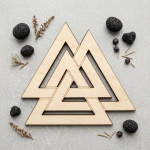 Op de afbeelding: Een lichtgekleurd houten Valknut-symbool bestaande uit drie in elkaar grijpende driehoeken. Het uitgesneden ontwerp is omringd door verschillende kleine, donkere, poreuze vulkanische stenen en takjes gedroogde heide op een neutrale ondergrond.