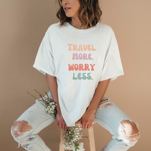 Könnte beinhalten: Ein weißes T-Shirt mit dem Text "TRAVEL MORE. WORRY LESS." in bunten, abgerundeten Buchstaben.