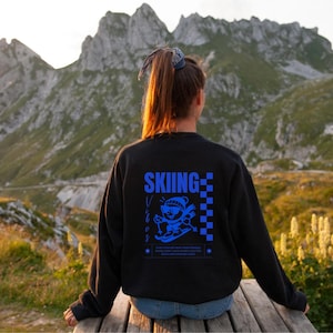 Könnte beinhalten: Schwarzes Sweatshirt mit einem blauen Aufdruck auf dem Rücken mit der Aufschrift "Skiing Vibes" und einem Cartoon-Skifahrer. Das Sweatshirt wird von einer Person getragen, die auf einer Holzbank mit Bergkulisse sitzt.