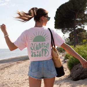 Op de afbeelding: Een roze T-shirt met een groene zon en de tekst "SONNEN KIND" op de rug. De persoon draagt ​​jeans shorts en loopt op een zandstrand.