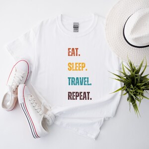 Könnte beinhalten: Weißes T-Shirt mit dem Text "EAT. SLEEP. TRAVEL. REPEAT." in bunten Retro-Lettern.
