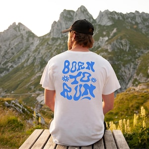 Op de afbeelding: Een wit T-shirt met een blauwe grafische afbeelding waarop "Born to Run" staat in een retro lettertype. Het shirt wordt gedragen door een persoon die voor een bergketen staat.
