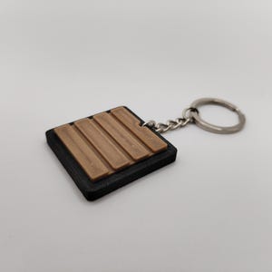 Könnte beinhalten: Ein quadratischer Schlüsselanhänger mit schwarzem Rand und braunem, strukturiertem Design. Der Schlüsselanhänger hat eine silberne Kette und einen Ring. Das Design scheint eine stilisierte Darstellung von Holzplanken zu sein.