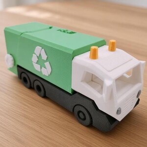 Peut inclure: Un camion poubelle jouet avec une carrosserie verte ornée d'un symbole de recyclage blanc, une cabine blanche, un châssis gris foncé avec des roues et deux petits feux jaunes sur le toit. Le camion a une surface texturée et un bouton blanc.