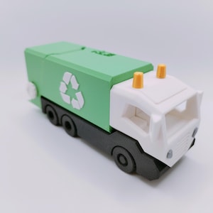 May include: A toy garbage truck with a green recycling bin and a white cab. The truck has black wheels and a recycling symbol on the side. Two orange lights are on top of the cab.