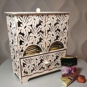 Puede incluir: Una caja de té de madera de color claro con intrincados recortes florales. La caja tiene un cajón y una abertura curva para las bolsitas de té. Una caja de "Spring Melody Black Tea" y bombones envueltos están al lado.