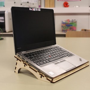 May include: A silver laptop computer is resting on a wooden stand. The laptop is open and the screen is black. The stand is made of wood and has a cutout design.