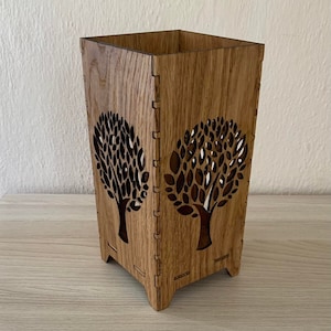 Puede incluir: Un portavelas de madera con un diseño de árbol recortado. El portavelas está hecho de madera marrón claro y tiene una forma cuadrada.