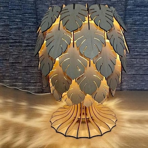 Op de afbeelding: Een houten tafellamp met een uniek ontwerp met een cluster van lasergesneden bladeren. De bladeren zijn in een cirkelvormige opstelling geplaatst en creëren een warme, uitnodigende gloed wanneer de lamp aan staat.