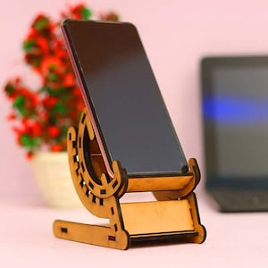 May include: A wooden phone stand with a unique design. The stand is made of light brown wood and has a curved shape. A black smartphone is placed in the stand.